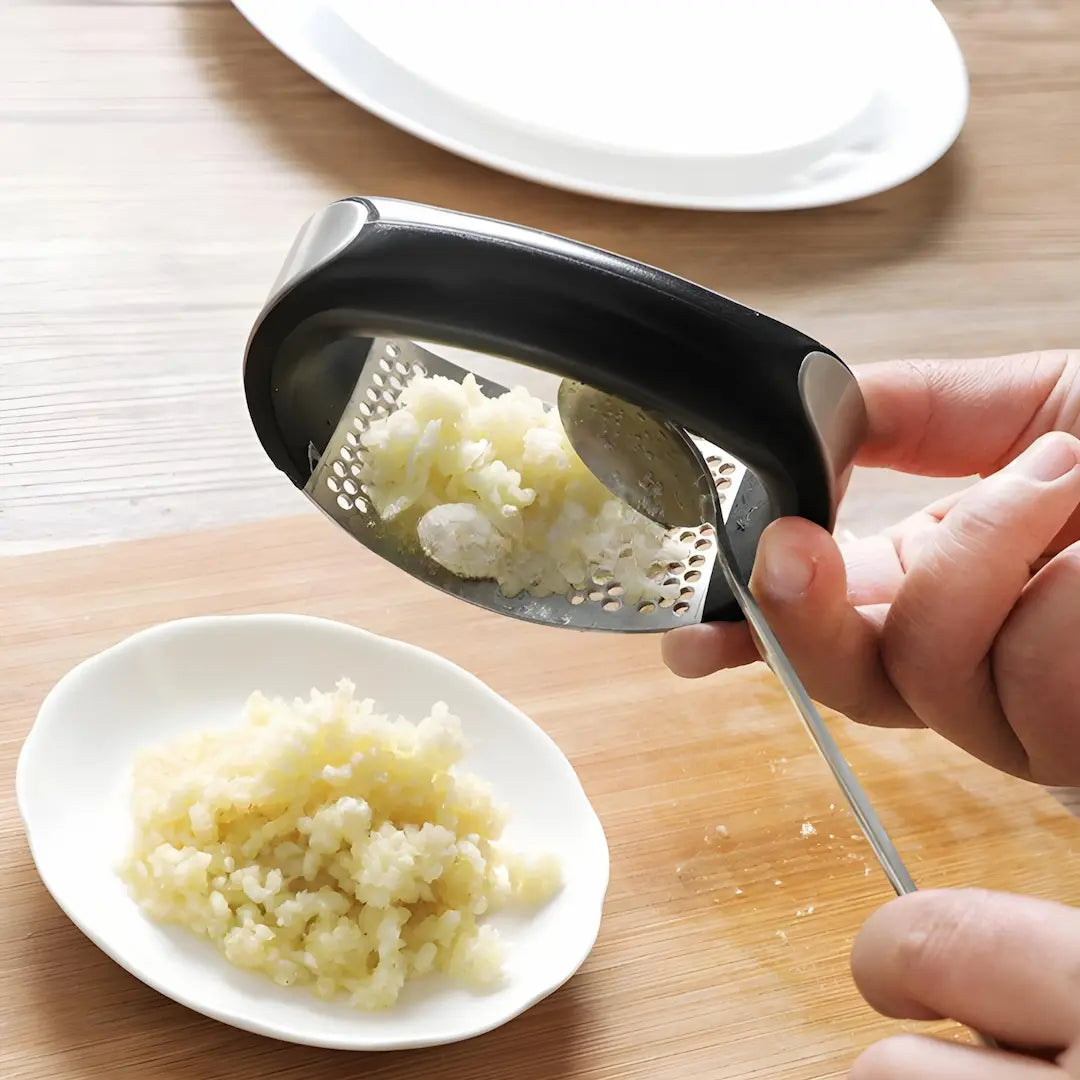 
A imagem mostra um amassador de alho em uso. Uma mão segura o utensílio, que possui design curvado e cabo preto, enquanto o alho triturado é transferido para um prato branco pequeno. A superfície de trabalho é de madeira, e há um prato maior ao fundo. O processo parece prático e eficiente.