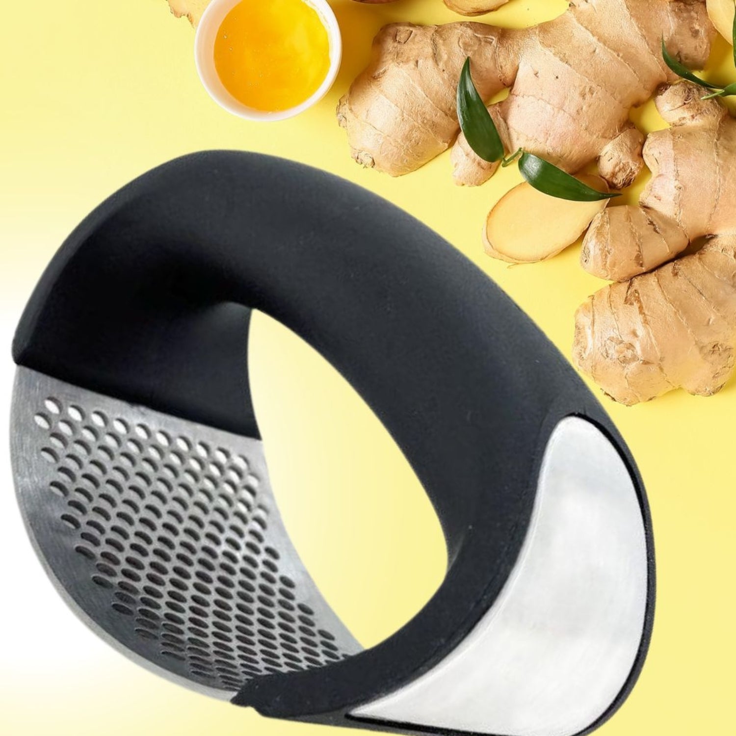 A imagem destaca um amassador de alho com design ergonômico e cabo preto curvado, acompanhado de uma superfície metálica perfurada para triturar alimentos. Ao fundo, sobre um fundo amarelo, há gengibres frescos cortados e uma pequena tigela com molho amarelo, sugerindo seu uso versátil.