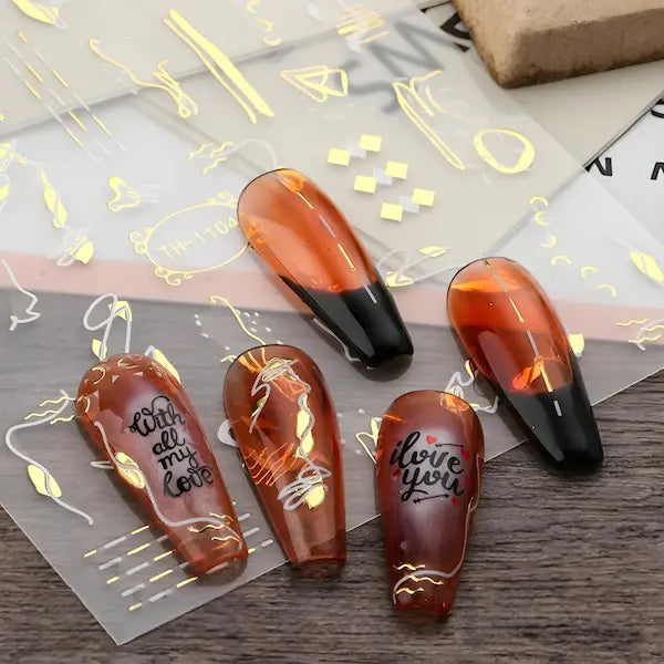 A imagem mostra cinco unhas postiças em tom marrom-alaranjado, duas com ponta preta e três com designs decorativos. As unhas possuem frases como "with all my love" e "I love you" em preto. Ao fundo, há folhas transparentes com decalques metálicos dourados em formas abstratas, prontos para serem aplicados nas unhas.