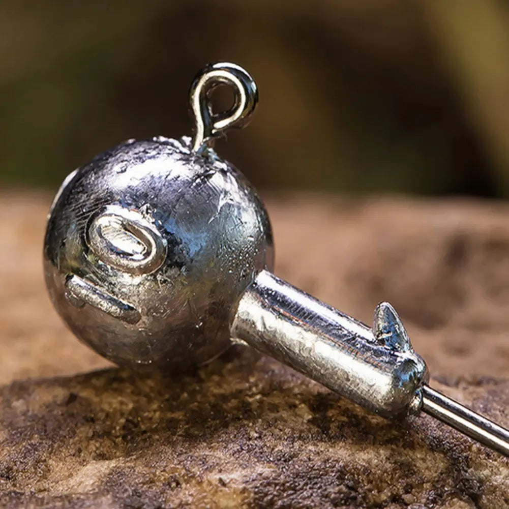 A imagem mostra um close-up de um anzol de pesca de aço inoxidável sobre uma superfície de pedra. O foco está na esfera metálica do anzol, que tem um laço no topo para prender a linha. A esfera é brilhante e possui marcas visíveis, indicando uma construção robusta.