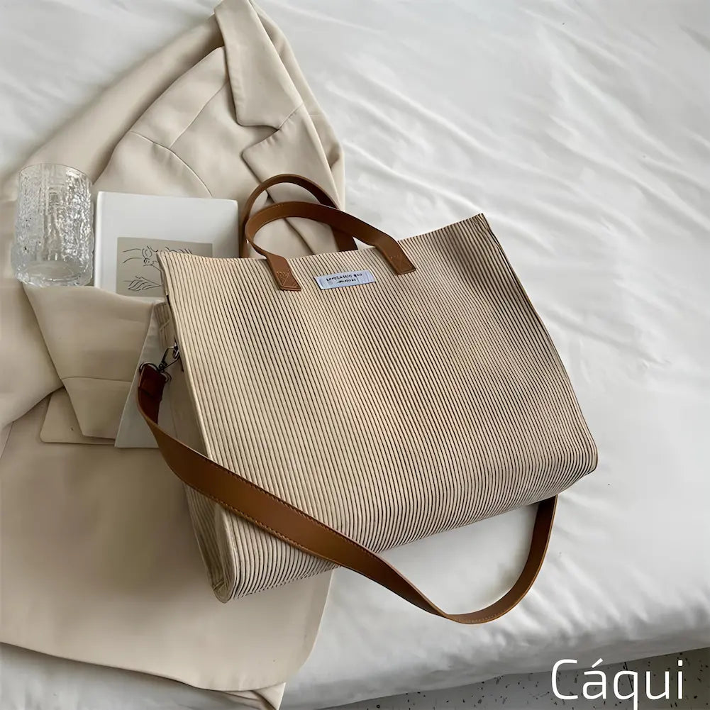 Imagem mostrando uma bolsa de cor cáqui. A bolsa está sobre uma cama branca, ao lado de um blazer bege, um copo de vidro e um caderno. A bolsa tem alças marrons e uma etiqueta branca. Esta é uma das opções de compra.