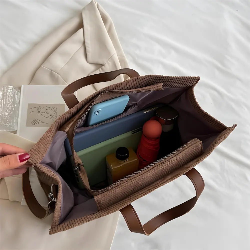 A imagem mostra uma bolsa marrom de tecido canelado aberta, revelando seu interior organizado. Dentro, há um telefone, laptop, caderno, garrafa, e outros itens pessoais. As alças são de couro sintético marrom claro. A bolsa está sobre um blazer bege, junto com um copo de vidro e um caderno, dispostos sobre uma cama branca.