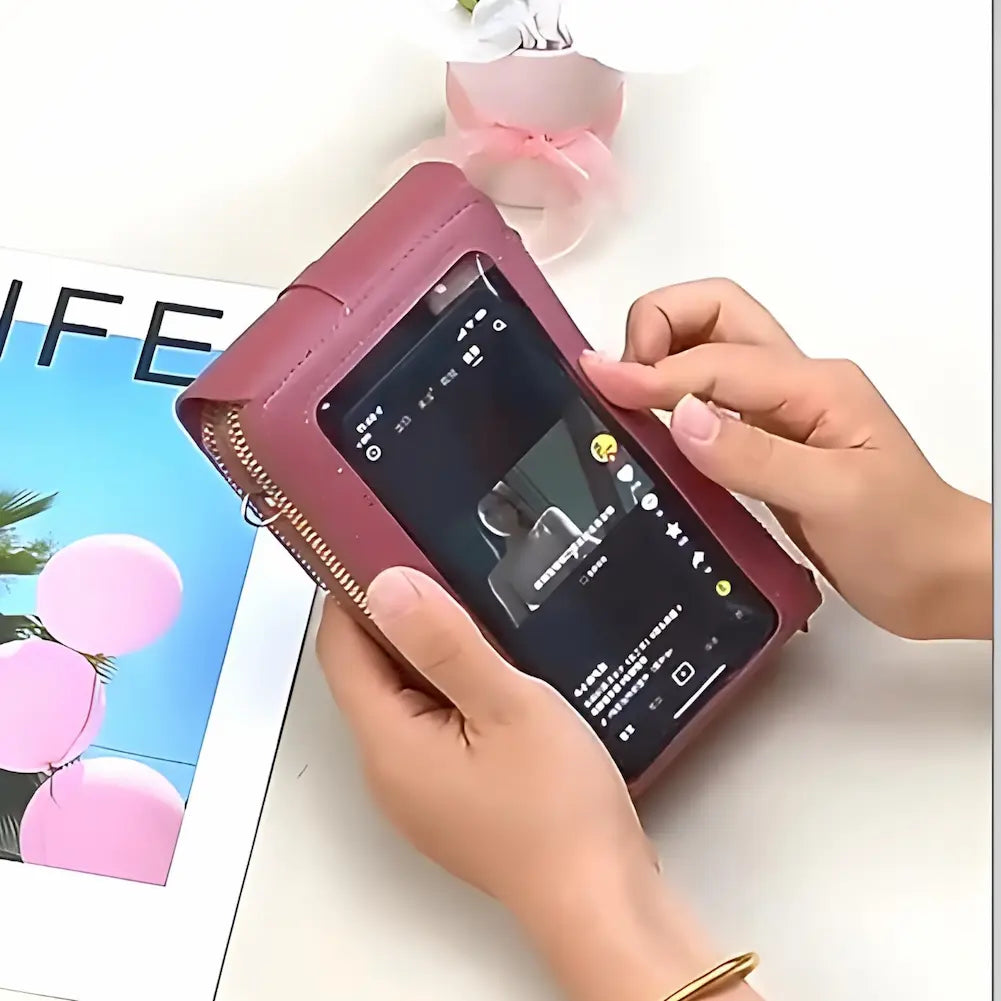 A imagem mostra uma pessoa usando um celular através de um visor transparente na parte traseira de uma bolsa tiracolo rosa da marca Endaycon. A bolsa está sendo segurada pelas mãos da pessoa e possui um zíper dourado. Ao lado, há uma revista com a capa que mostra balões cor-de-rosa e a palavra "LIFE", e uma pequena planta com flores brancas e fitas cor-de-rosa.