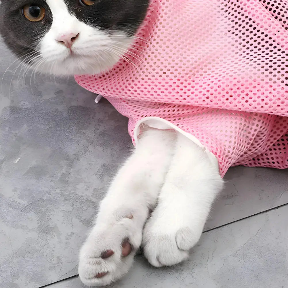  A imagem mostra um gato branco e preto deitado no chão, parcialmente dentro de uma bolsa de rede rosa. O gato olha diretamente para a câmera com uma expressão calma. As patas dianteiras do gato estão para fora da bolsa, descansando no chão cinza. A bolsa de rede cobre o corpo do gato, deixando apenas a cabeça e as patas visíveis.