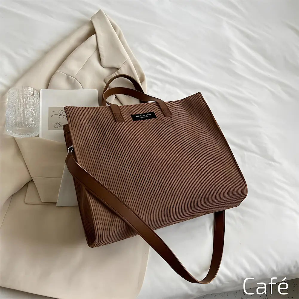 Imagem mostrando uma bolsa na cor café. Está sobre uma cama branca, acompanhada de um blazer bege, copo de vidro e caderno. A bolsa tem alças marrons e uma etiqueta preta. Esta é uma das opções de compra.