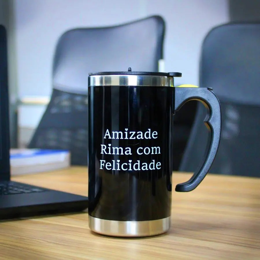 A imagem mostra uma caneca preta com detalhes em inox na parte inferior e uma tampa preta. A caneca possui a mensagem "Amizade Rima com Felicidade" escrita em branco, transmitindo uma mensagem positiva e alegre. Ela está posicionada sobre uma mesa de escritório, ao lado de um laptop, sugerindo que é uma opção ideal para quem gosta de desfrutar de bebidas quentes enquanto trabalha. O design é moderno e prático, adequado para o uso diário.