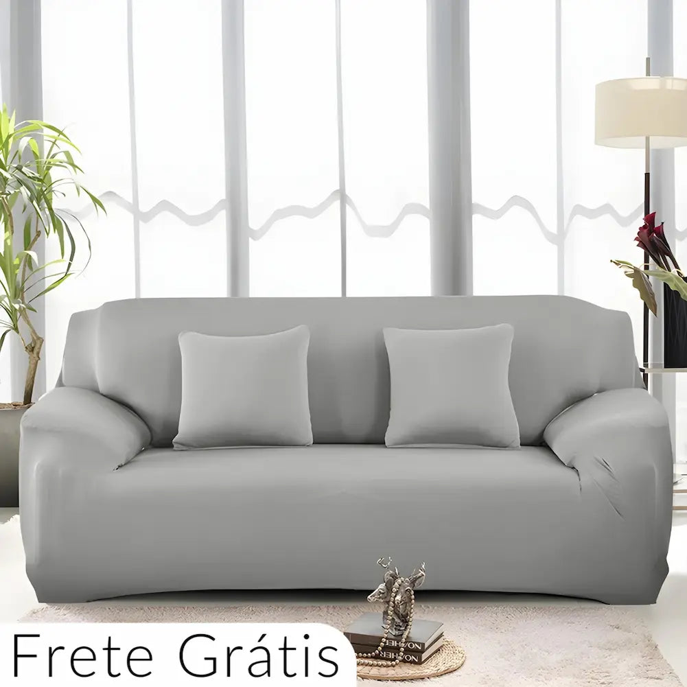  Imagem de uma sala clara com um sofá cinza claro. O sofá tem três almofadas cinza combinando. Ao lado esquerdo, há uma planta, e à direita, uma luminária de chão. Texto no canto inferior esquerdo: "Frete Grátis". Uma pequena mesa de centro com livros e uma escultura está à frente do sofá.