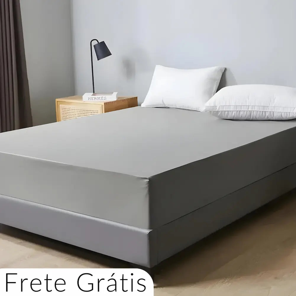  Imagem de uma cama com colchão coberto por uma capa cinza bem ajustada. A cama tem uma base cinza escura, dois travesseiros brancos e uma parede cinza ao fundo. Um abajur preto está sobre um criado-mudo ao lado. No canto inferior esquerdo, o texto diz "Frete Grátis".