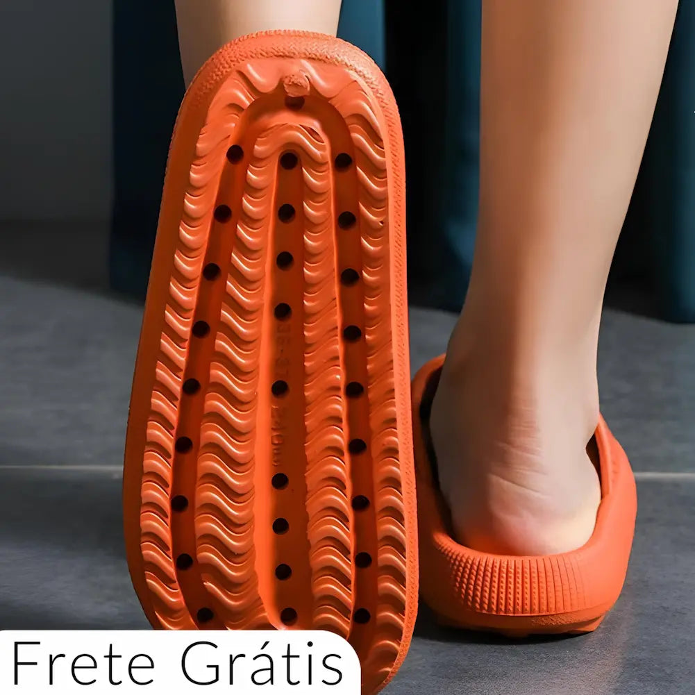 Imagem mostrando uma pessoa usando chinelos de banho laranja, destacando a sola do chinelo. A sola possui um design com ranhuras e furos para melhor tração e drenagem. O texto "Frete Grátis" aparece no canto inferior esquerdo, indicando que o produto é enviado sem custo adicional. A imagem enfatiza a segurança e praticidade dos chinelos, ideal para uso em ambientes molhados.