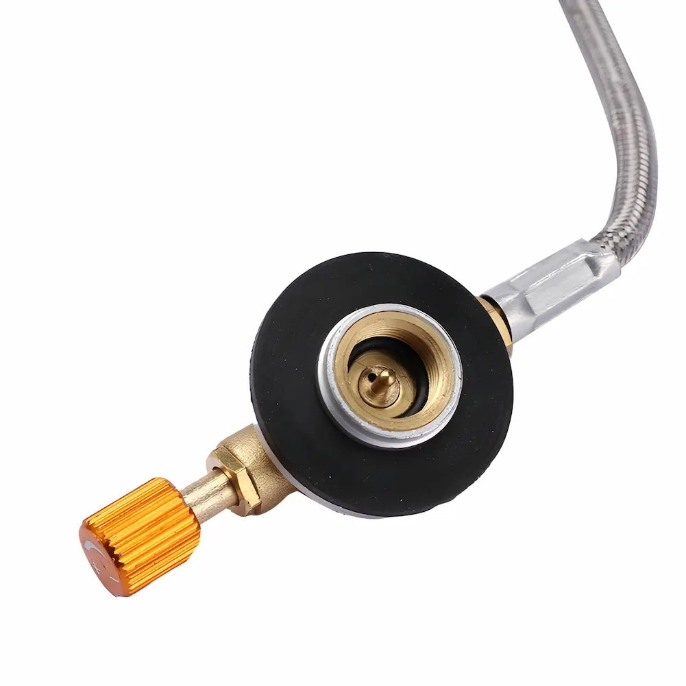 A imagem mostra um close-up da válvula de conexão de um fogareiro portátil. A válvula é feita de latão com uma base preta e uma ponta laranja, conectada a uma mangueira metálica. A válvula parece ser o ponto de controle do gás para o fogareiro.