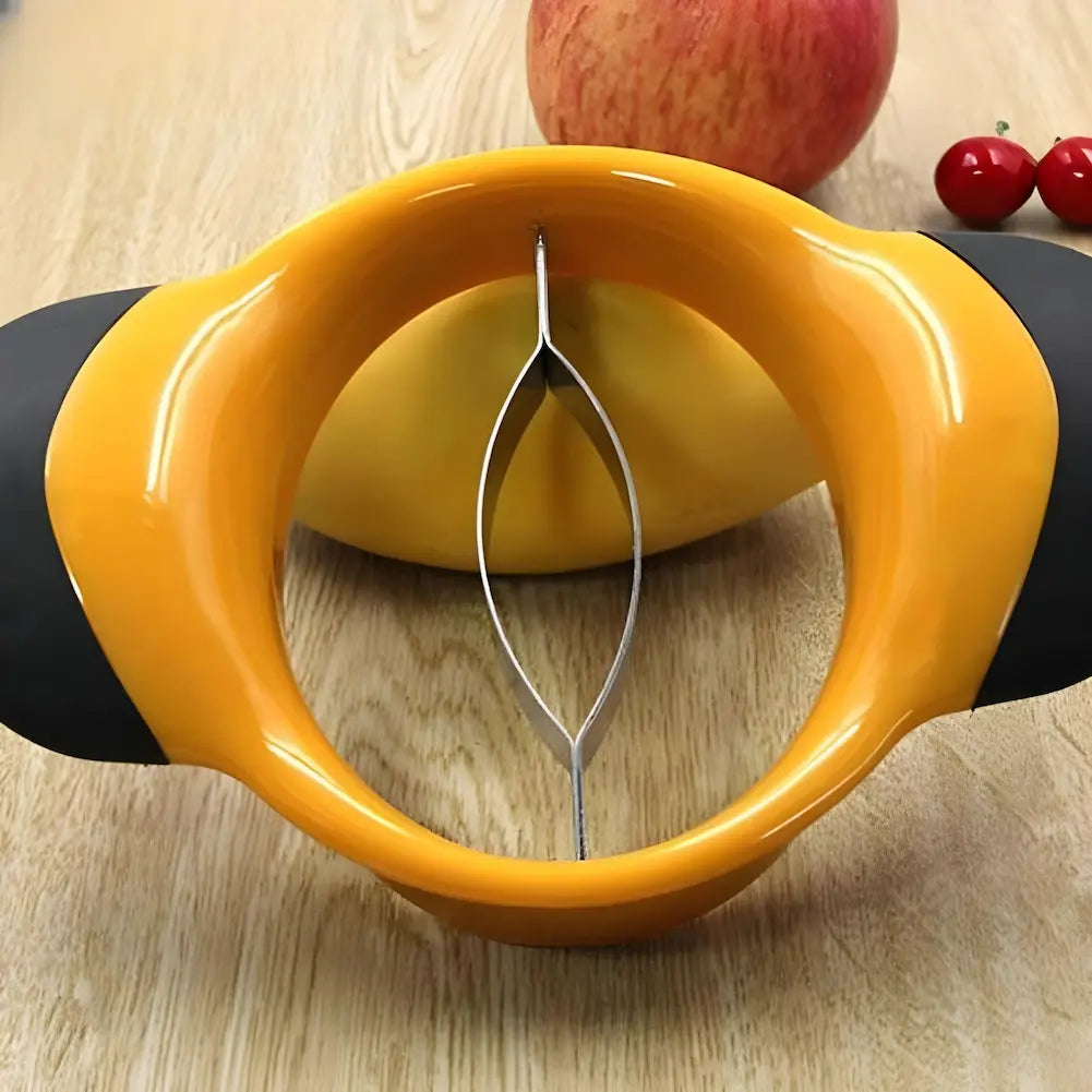 A imagem mostra um descascador de frutas amarelo com alças pretas, visto de frente. No centro, há uma lâmina curva de metal. O descascador está sobre uma superfície de madeira clara. Ao fundo, há uma manga, uma maçã e algumas cerejas decorativas.