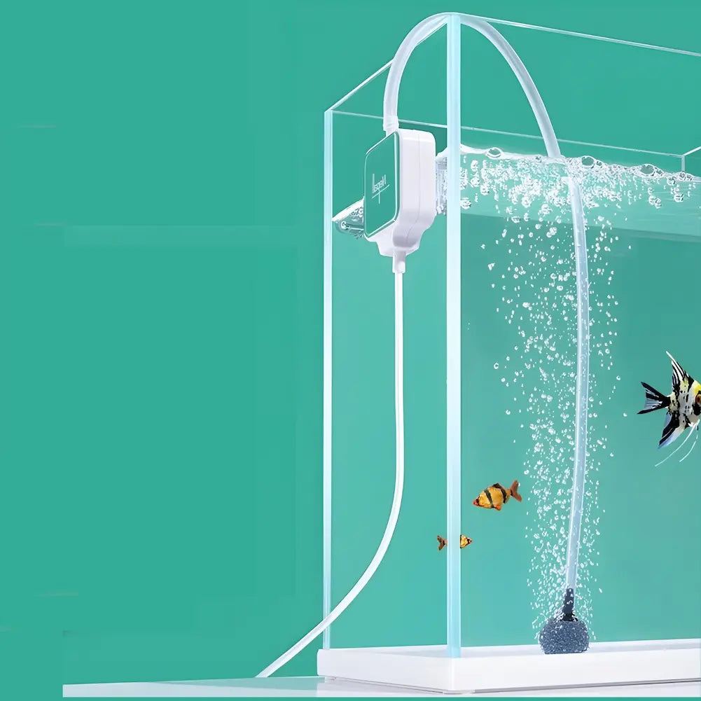 A imagem mostra um aquário com um dispositivo verde e branco da marca "Nerall" preso ao lado do vidro. Uma mangueira sai do dispositivo e conecta-se a uma pedra difusora no fundo do aquário, liberando bolhas de ar. Dentro do aquário, há alguns peixes coloridos nadando. O fundo é verde claro, destacando a estética limpa e moderna do aquário e do dispositivo.