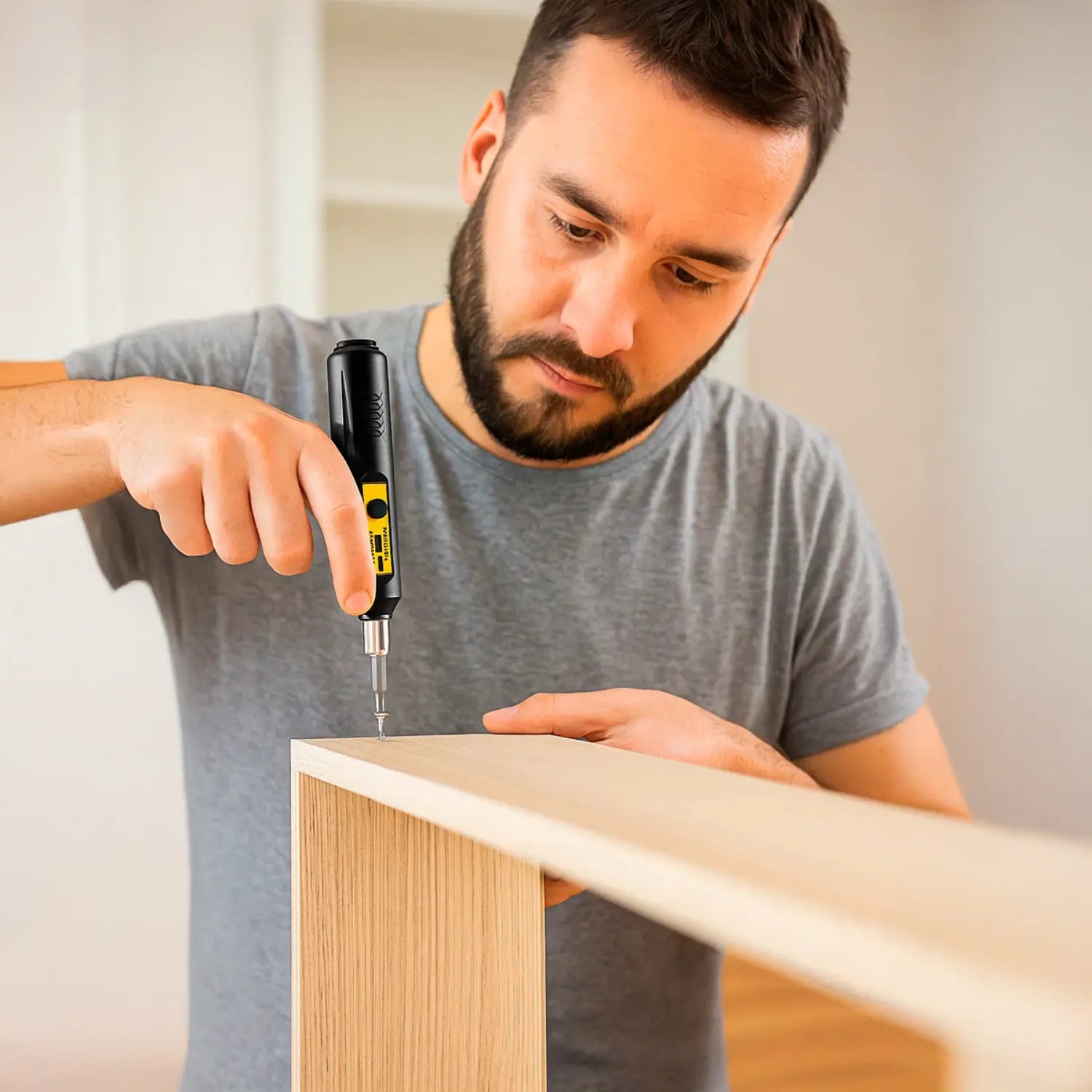 A imagem mostra um homem concentrado, usando uma parafusadeira elétrica para montar ou ajustar um móvel de madeira. Ele está com uma camiseta cinza e se encontra em um ambiente bem iluminado e moderno, com um estilo minimalista e clean. O foco está no trabalho cuidadoso de encaixar a peça de madeira, evidenciando a atenção e precisão necessárias para o processo de montagem.