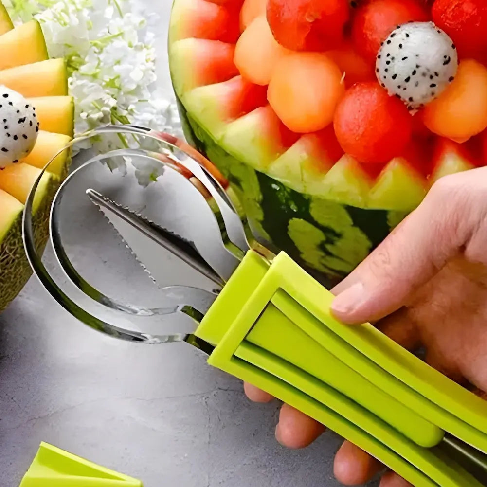  Na imagem, uma mão segura um utensílio de esculpir frutas com cabo verde e lâmina curva. Ao fundo, há uma melancia decorada com bolas de frutas coloridas e um melão cortado em forma de flor. Flores brancas estão ao lado, complementando a apresentação das frutas.
