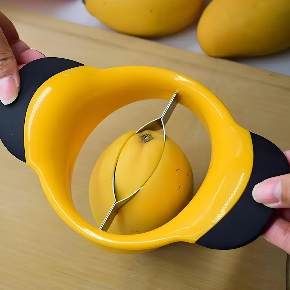 A imagem mostra uma pessoa usando um descascador de frutas amarelo com alças pretas para cortar uma manga. O descascador está posicionado sobre a manga, com a lâmina curva de metal no centro, começando a dividir a fruta. Ao fundo, há outras mangas sobre uma superfície de madeira clara.