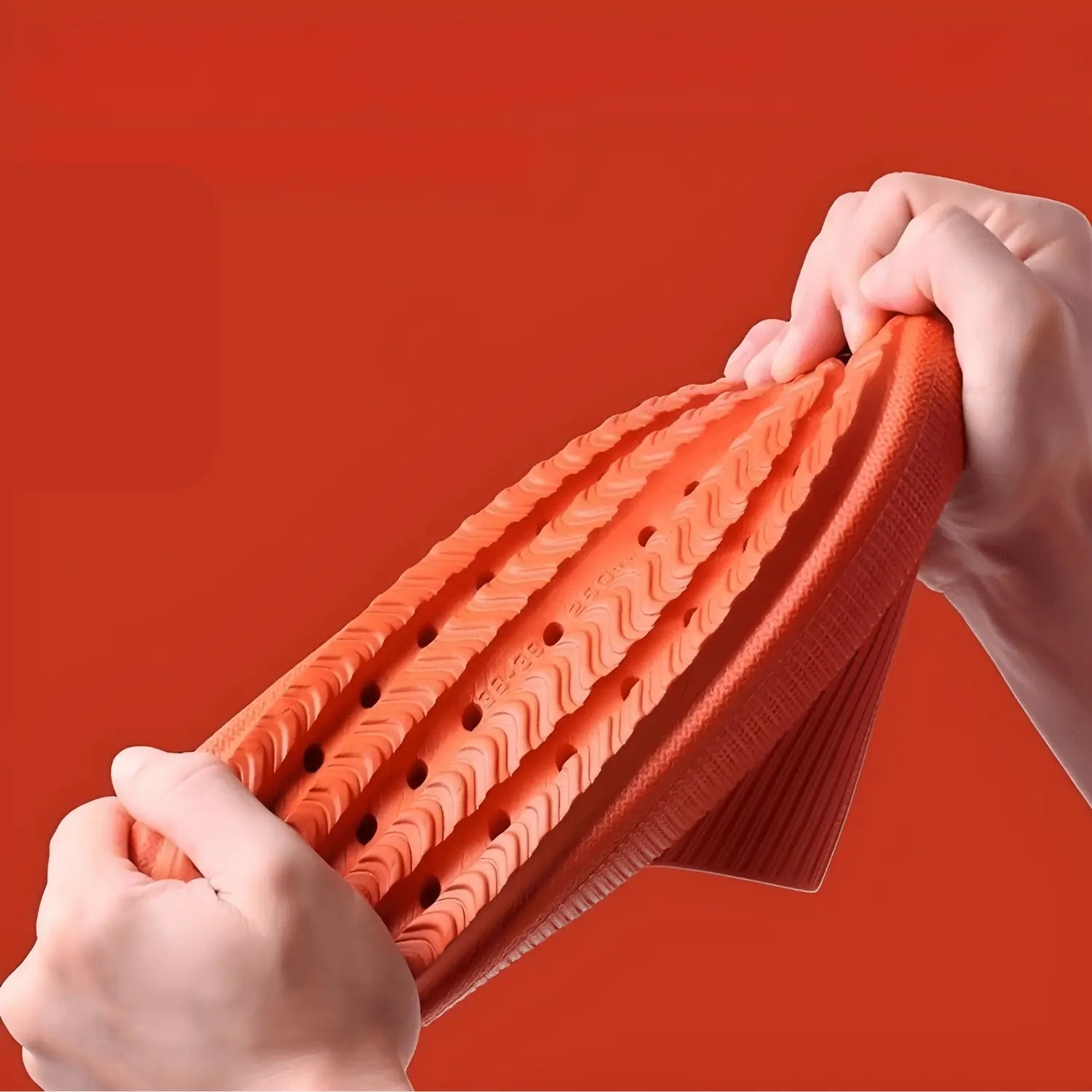 Imagem mostrando uma pessoa puxando e flexionando a sola de um chinelo de banho laranja para demonstrar sua flexibilidade e durabilidade. A sola possui ranhuras e furos para melhor tração e drenagem. O fundo laranja realça o material elástico e a construção robusta do chinelo, destacando sua qualidade e resistência.