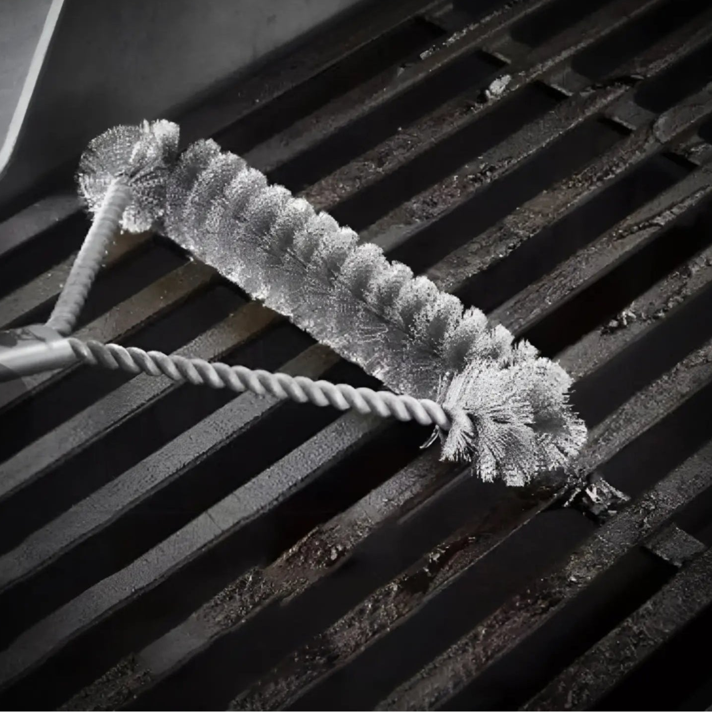 A imagem mostra uma escova para limpeza de grelha, com um design robusto e eficaz. Ela possui um cabo de metal torcido e uma cabeça com cerdas espessas dispostas em várias direções, permitindo uma limpeza profunda das superfícies da grelha. O produto está sendo mostrado em cima das barras da grelha, com foco nas cerdas que entram em contato com a sujeira acumulada.