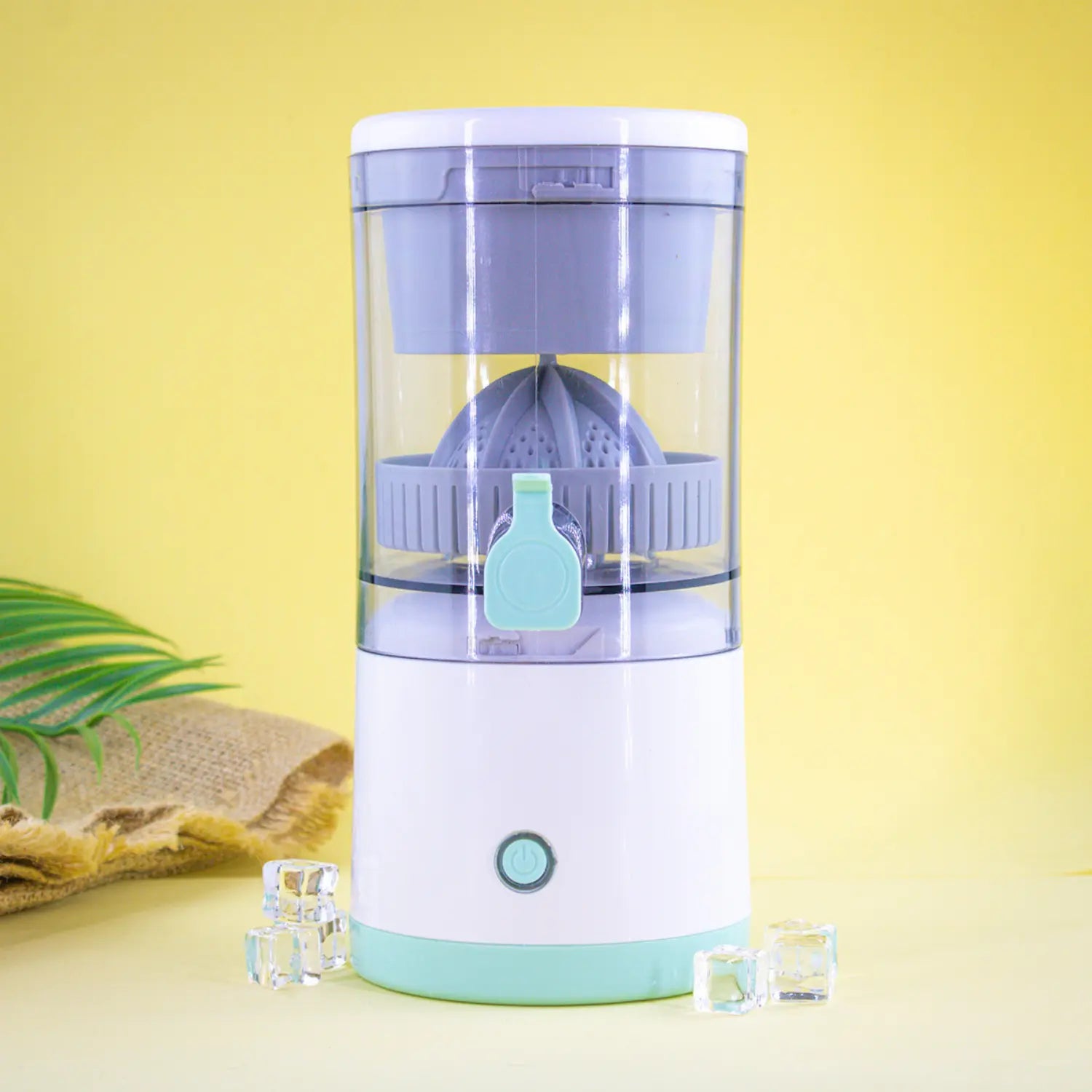 A imagem apresenta um espremedor de frutas moderno, com design compacto e cores suaves. A base é branca, com detalhes em verde claro, e a parte superior é transparente, permitindo visualizar o interior. O espremedor possui um compartimento para frutas, um botão de operação na base e está decorado com cubos de gelo ao lado. O fundo é amarelo, criando um ambiente alegre e vibrante.