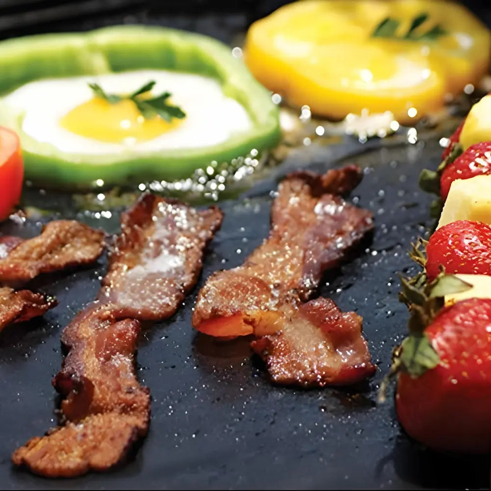  A imagem mostra uma variedade de alimentos sendo grelhados em um tapete antiaderente preto. Há tiras de bacon, fatias de pimentão com ovos no centro, e espetinhos de frutas com morangos e pedaços de queijo. Os alimentos estão dispostos de forma organizada e cozinhando uniformemente.