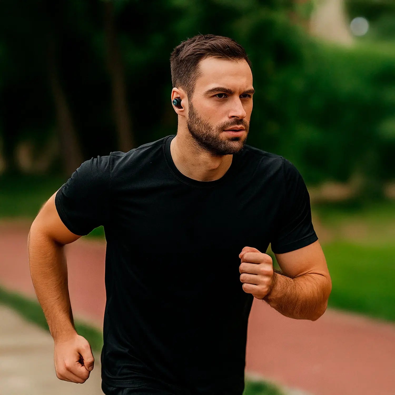 A imagem mostra um homem correndo ao ar livre enquanto usa fones de ouvido sem fio. Ele está vestindo uma camiseta preta e aparenta estar concentrado na corrida. Os fones de ouvido são pequenos e discretos, proporcionando conforto e praticidade durante a atividade física. O ambiente ao fundo é ao ar livre, com uma pista de corrida visível, destacando a ideia de que os fones são ideais para esportes e exercícios.