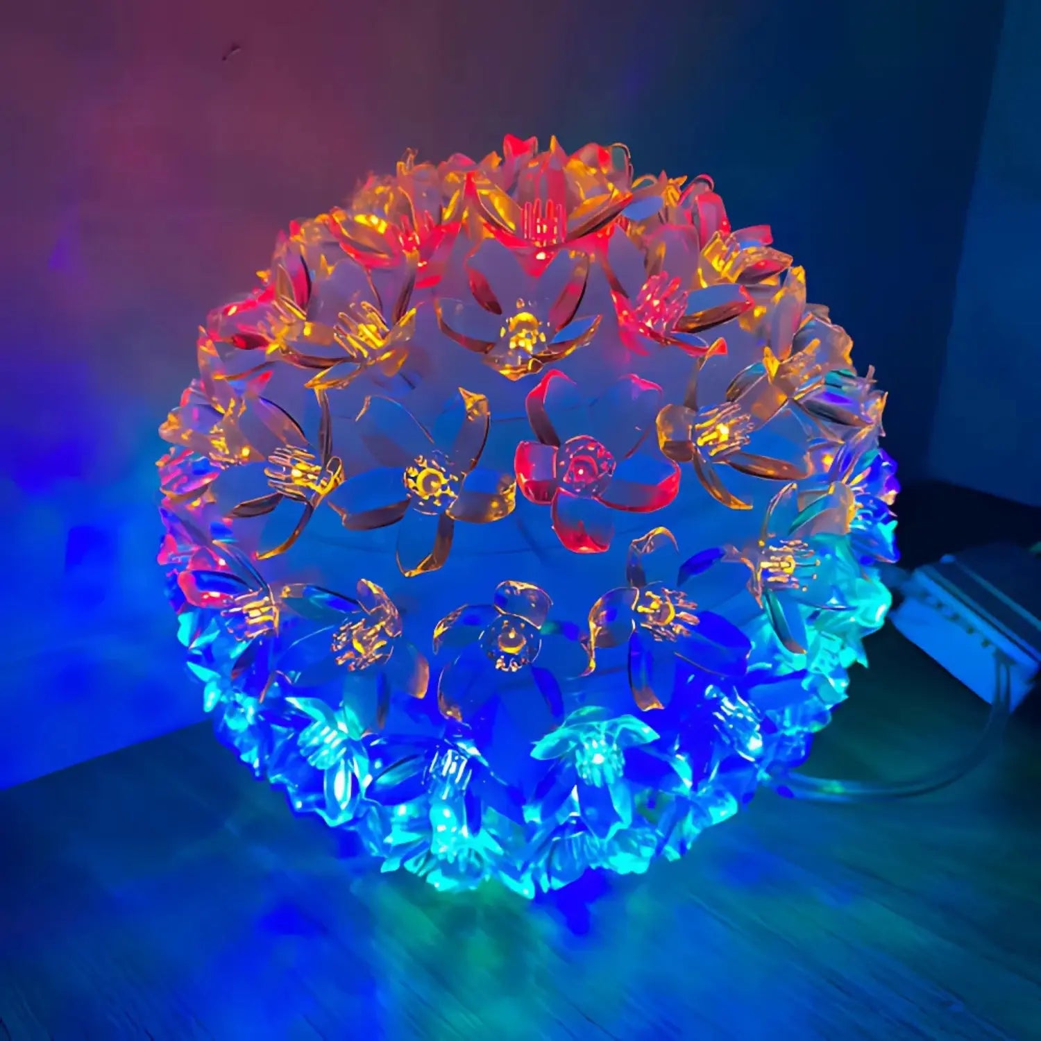 A imagem mostra uma luminária decorativa em forma de esfera, composta por flores feitas de material translúcido e iluminadas por luzes coloridas. As flores emitem tons de azul, vermelho e amarelo, criando um efeito visual vibrante e bonito, ideal para ambientes de decoração com um toque moderno e acolhedor.