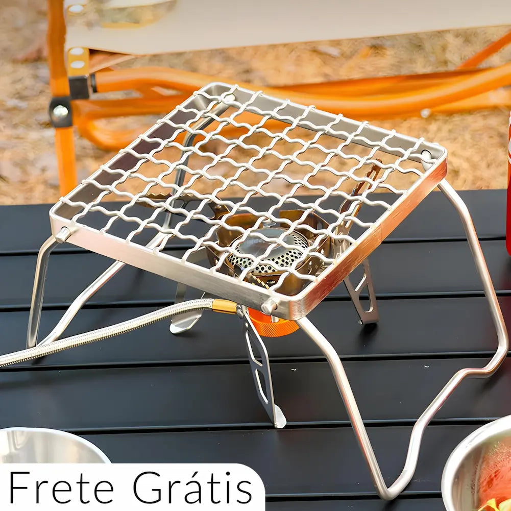 Imagem de uma grelha em aço dobrável para mini fogão, posicionada sobre uma mesa de acampamento. A grelha é quadrada, com uma estrutura metálica robusta e está conectada a um tubo flexível de gás. A imagem mostra o ambiente de acampamento ao fundo e inclui um texto "Frete Grátis" na parte inferior.