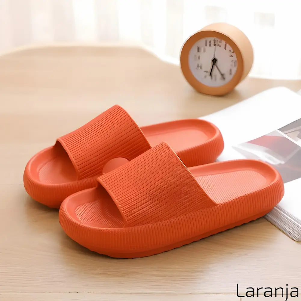 Imagem mostrando um par de chinelos laranja. Eles estão dispostos sobre uma superfície de madeira clara, ao lado de um relógio de mesa laranja e uma revista aberta, com iluminação natural suave ao fundo.