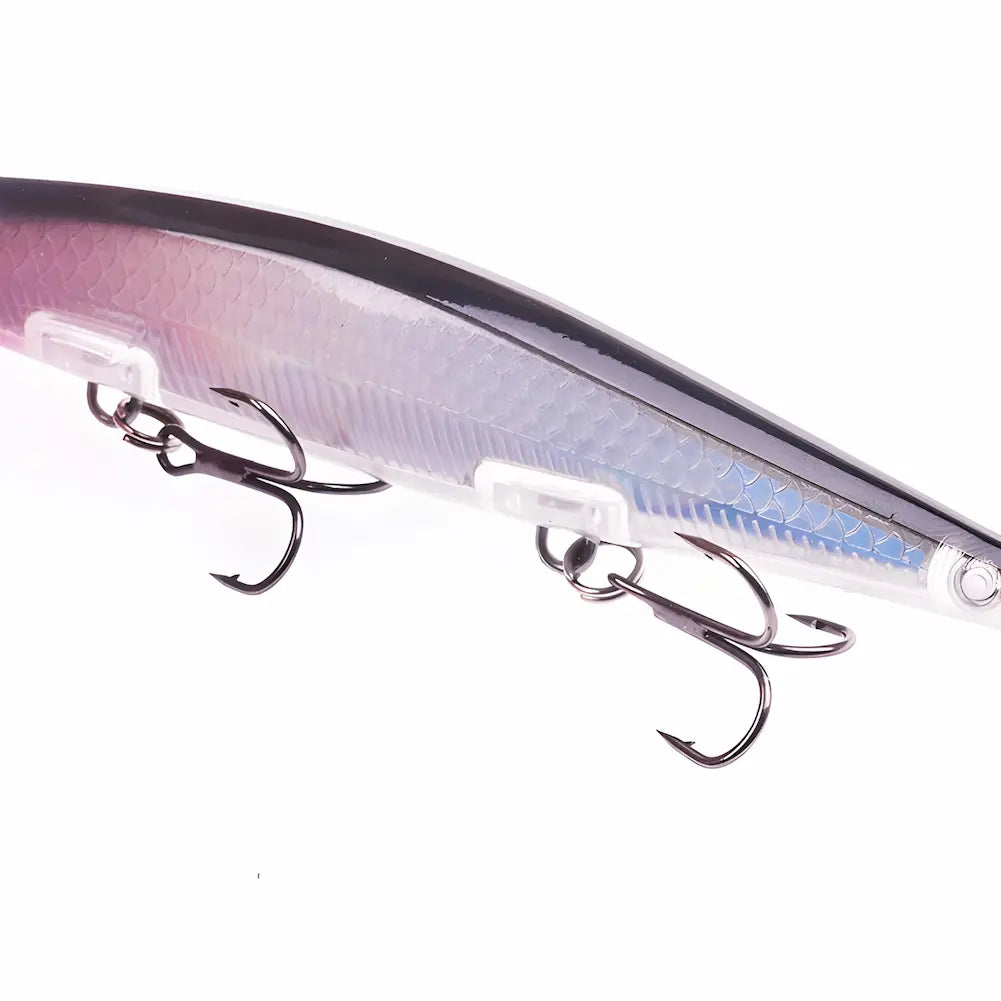 A imagem mostra uma isca de pesca artificial com corpo alongado e superfície texturizada. A isca é predominantemente prateada com tons de rosa e azul, além de detalhes pretos na parte superior. Possui dois anzóis triplos de cor escura. O fundo é branco, destacando os detalhes da isca.