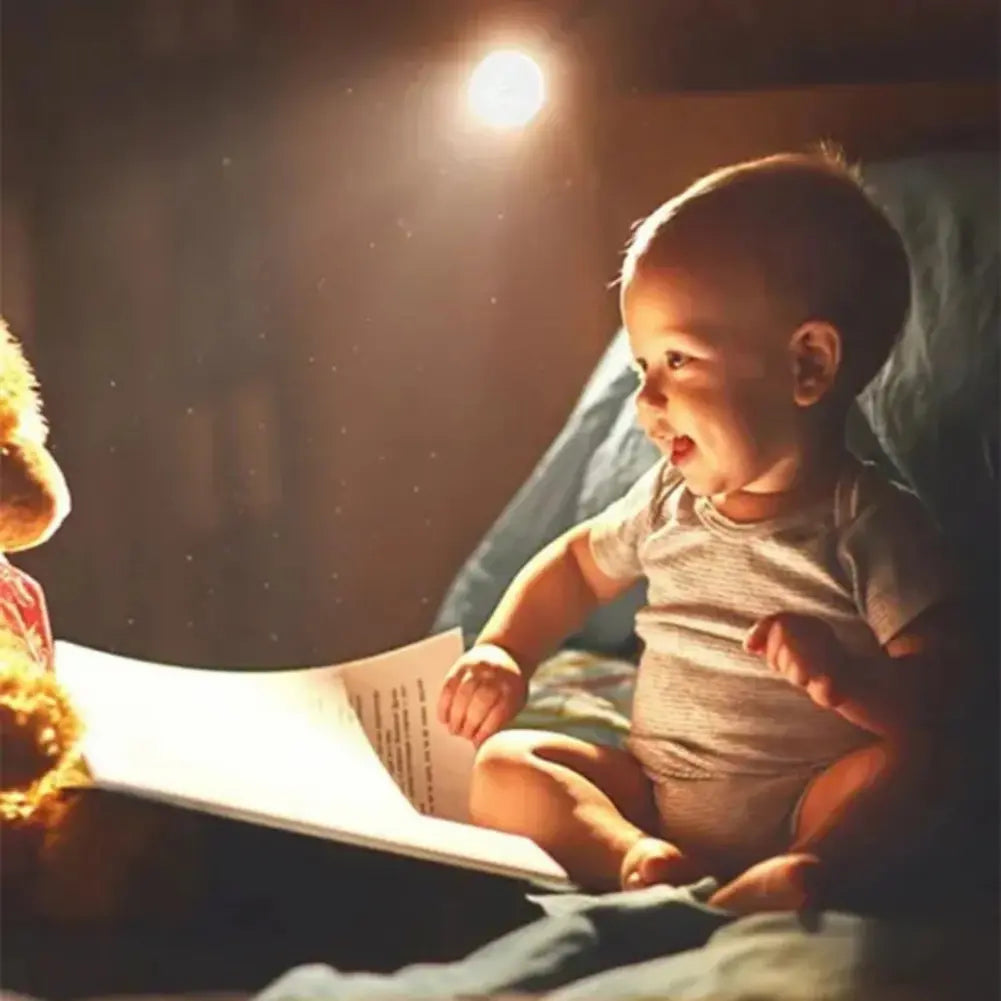  Imagem de um bebê sentado em uma cama, olhando animado para um livro grande aberto. Um ursinho de pelúcia está ao lado do livro. Uma luminária redonda de LED ilumina suavemente a cena, criando um ambiente acolhedor.