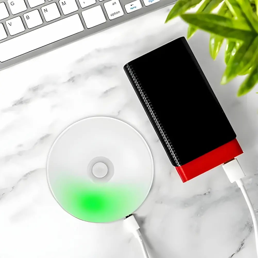  Imagem de uma luminária redonda conectada a um carregador USB preto e vermelho, ambos sobre uma superfície branca. A luminária está parcialmente iluminada em verde. Acima, parte de um teclado de computador é visível. Há uma planta verde no canto superior direito.