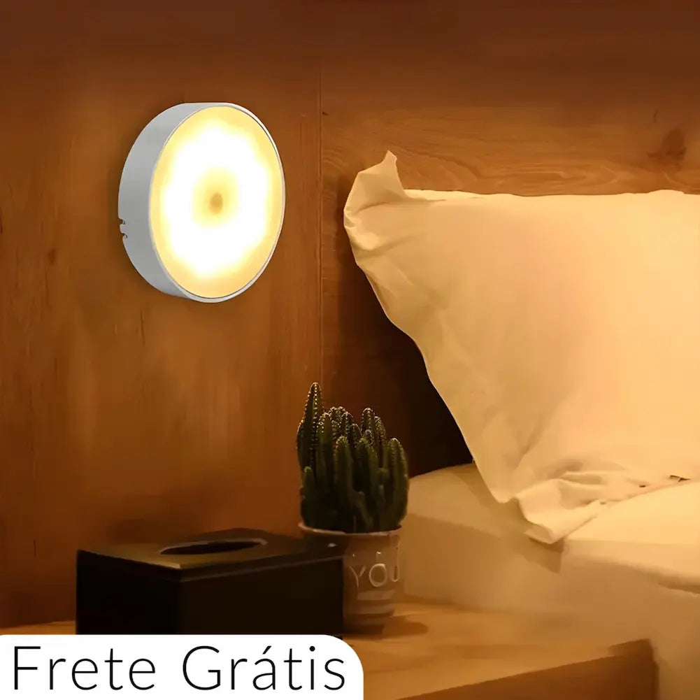  Imagem de uma luminária redonda com luz amarela suave fixada na parede ao lado de uma cama com travesseiro branco. Abaixo, há uma mesa de cabeceira com um cacto em um vaso e uma caixa preta. Texto na parte inferior diz "Frete Grátis".