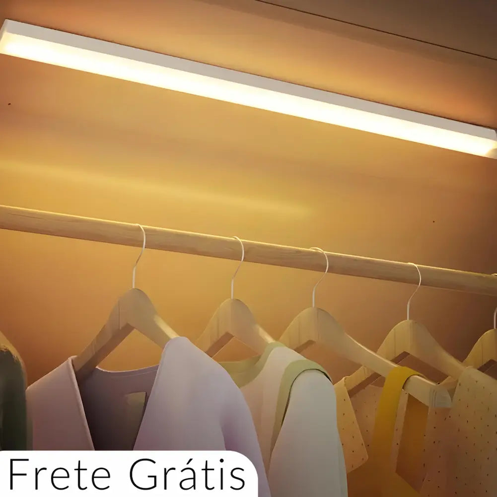  A imagem mostra uma luminária retangular emitindo luz quente, instalada na parte superior de um guarda-roupa. Abaixo da luminária, há um varão com cabides de madeira pendurados, segurando roupas. No canto inferior esquerdo, há um texto que diz "Frete Grátis".