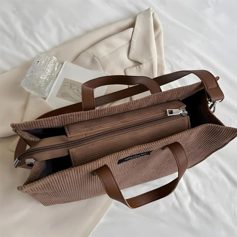 A imagem mostra uma bolsa marrom de tecido canelado, aberta, revelando seu interior espaçoso com vários compartimentos. As alças são de couro sintético marrom claro. A bolsa está sobre um blazer bege, junto com um copo de vidro e um caderno, todos dispostos sobre uma cama branca.