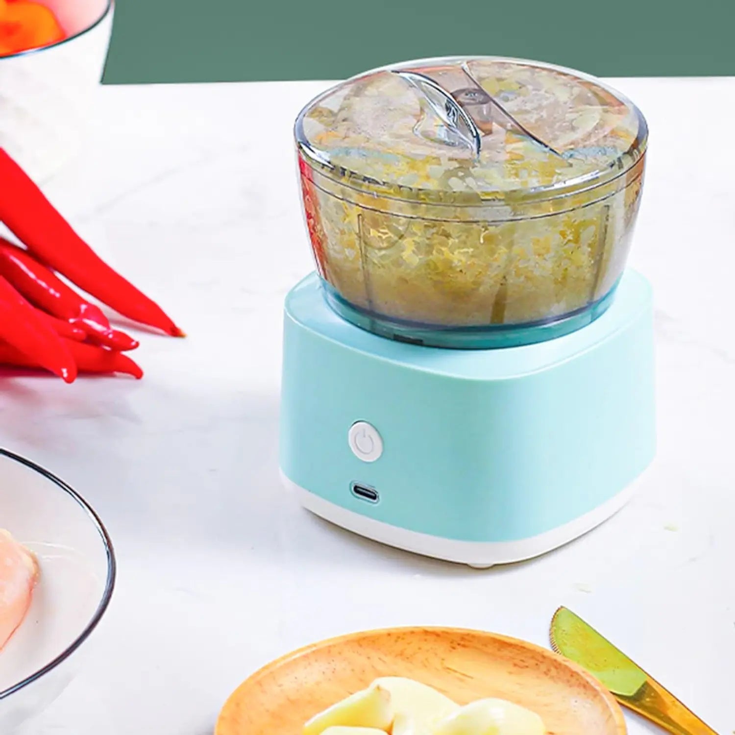 A imagem mostra um processador de alimentos compacto, com um design moderno em azul. O recipiente transparente permite visualizar o conteúdo triturado, enquanto a base tem um botão de controle para acionar o aparelho. Ao redor, há ingredientes como pimentas e temperos, sugerindo o uso do processador para picar e misturar diversos alimentos de forma rápida e eficiente na cozinha.