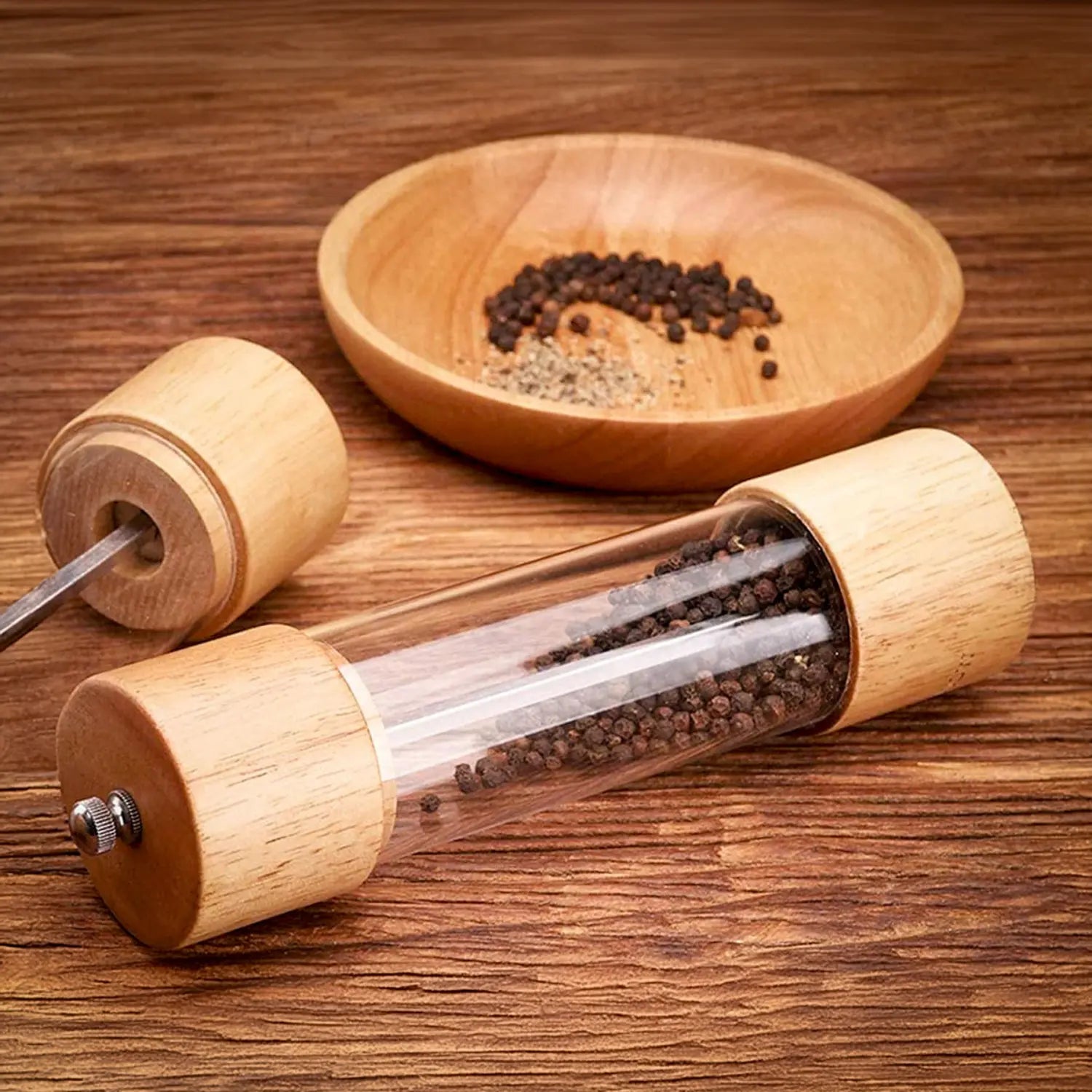 A imagem mostra um moedor de pimenta elegante com corpo de madeira e uma seção central de vidro, permitindo ver os grãos de pimenta dentro. O moedor está ao lado de um prato de madeira com pimenta moída, demonstrando sua funcionalidade. O design simples e natural confere um toque rústico e sofisticado, ideal para temperar alimentos de forma prática e charmosa na cozinha.