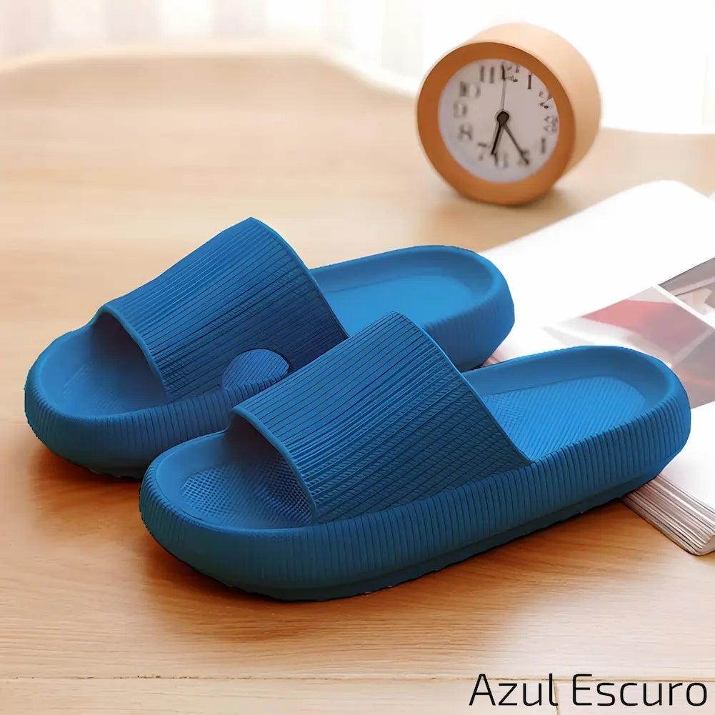 Imagem mostrando um par de chinelos azul escuro. Eles estão dispostos sobre uma superfície de madeira clara, ao lado de um relógio de mesa laranja e uma revista aberta, com um ambiente iluminado naturalmente.