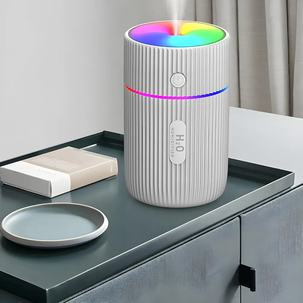 Imagem de um umidificador de ar portátil branco com textura ondulada, colocado sobre uma superfície cinza escura. O topo do umidificador emite luzes coloridas em arco-íris, e há um anel luminoso ao redor do meio do aparelho. Ao lado do umidificador, há um pequeno prato cinza e uma embalagem de cor clara. Ao fundo, há uma cortina bege.