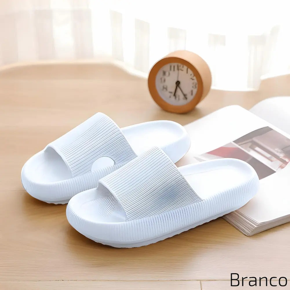 Imagem mostrando um par de chinelos brancos. Eles estão dispostos sobre uma superfície de madeira clara, ao lado de um relógio de mesa laranja e uma revista aberta, em um ambiente minimalista e elegante.