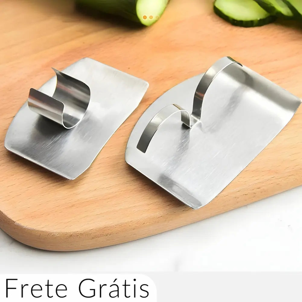A imagem mostra duas lâminas curvas de metal sobre uma tábua de corte de madeira clara. Cada lâmina tem um suporte de metal para os dedos. Ao fundo, há fatias de pepino. Na parte inferior da imagem, está escrito "Frete Grátis" em texto preto sobre um fundo branco.
