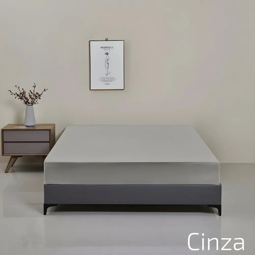  A imagem mostra um quarto minimalista. No centro, há uma cama com um protetor de colchão cinza. Ao lado da cama, há uma mesa de cabeceira com uma base de madeira e duas gavetas cinza. Sobre a mesa, uma jarra branca com galhos secos. Na parede, um quadro simples com uma ilustração de uma pessoa.