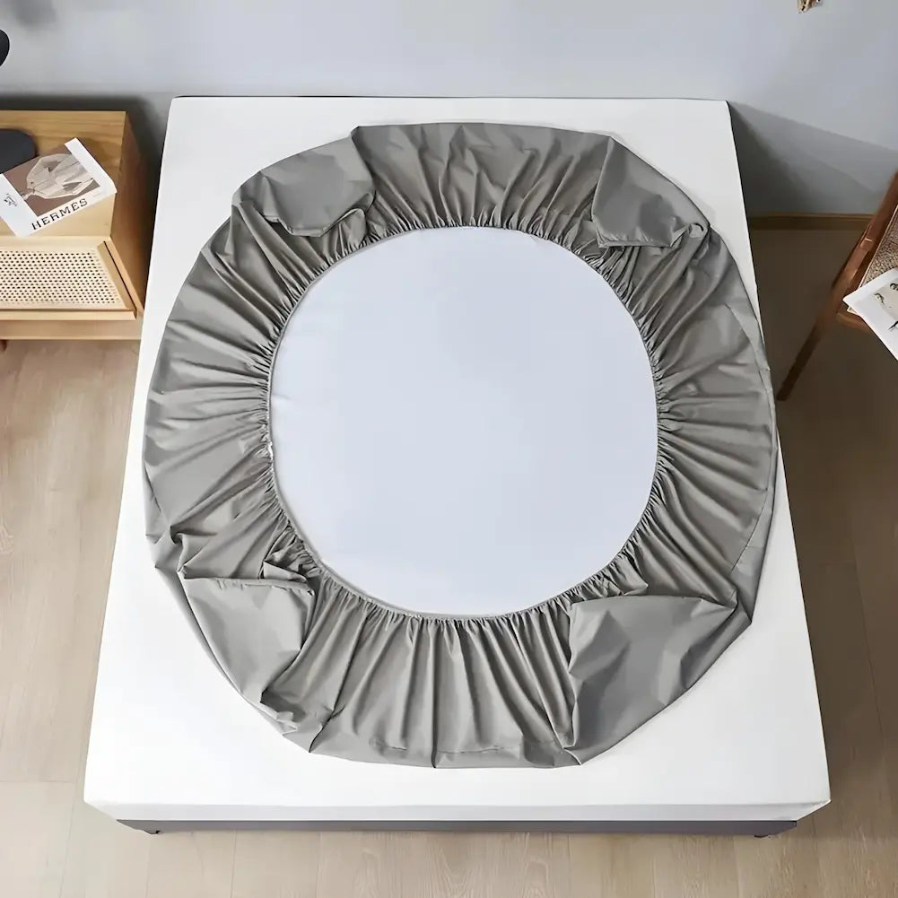 Imagem de uma cama vista de cima com um colchão branco. Sobre o colchão, uma capa de colchão cinza claro, ainda não totalmente ajustada, com bordas elásticas ao redor. A capa forma um círculo solto no centro do colchão. Ao lado da cama, um criado-mudo com um livro e uma luminária.