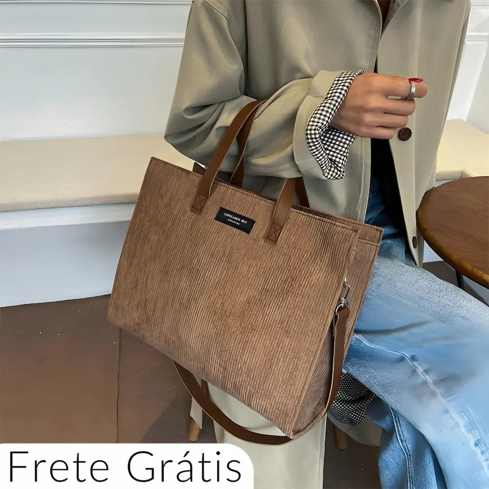 A imagem mostra uma bolsa marrom texturizada com alças duplas e um detalhe preto com o texto "LIFELAND BAG". Uma pessoa vestida com um casaco bege e jeans segura a bolsa. No canto inferior esquerdo, há um texto dizendo "Frete Grátis".