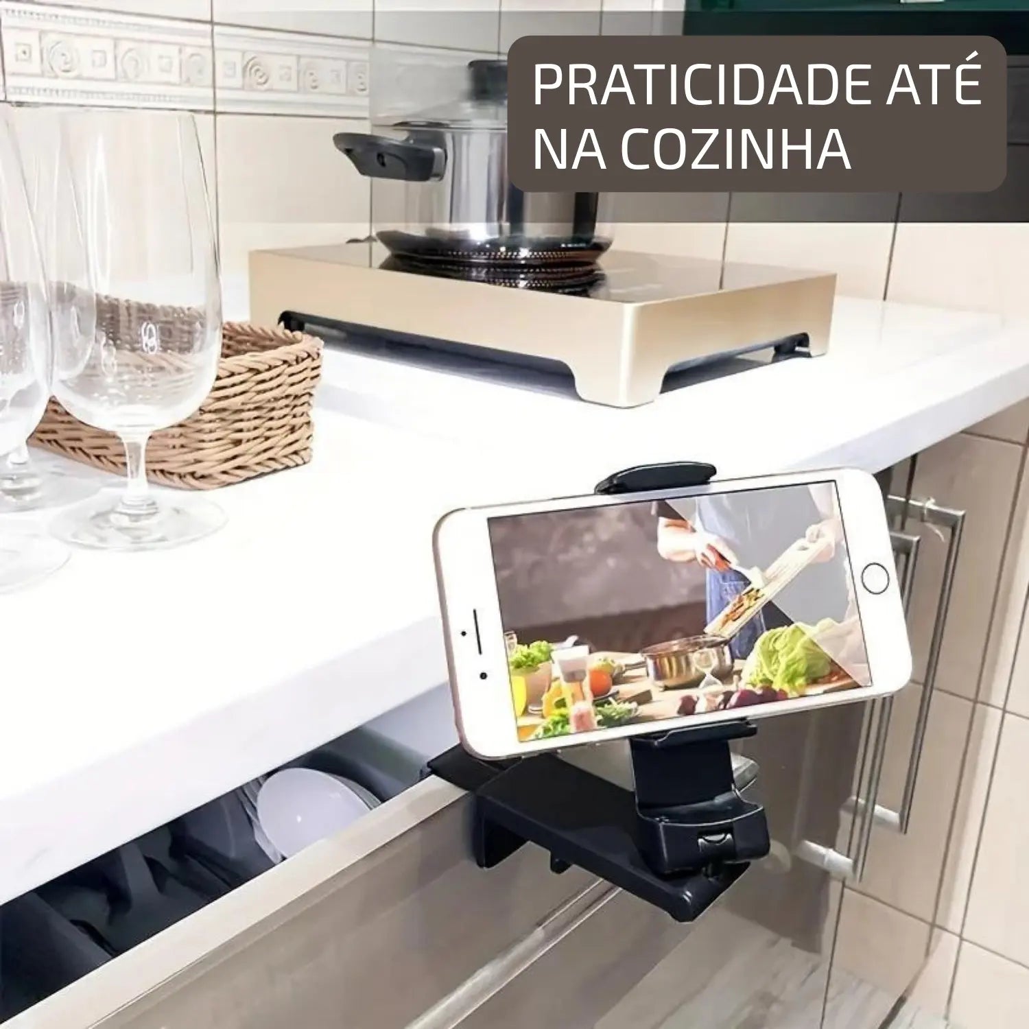 A imagem mostra um suporte de celular preso a uma gaveta de cozinha, com o celular exibindo um vídeo, possivelmente relacionado a uma receita. Ao fundo, há um fogão e utensílios de cozinha, criando um ambiente prático para quem está cozinhando e precisa de instruções ou receitas enquanto usa as mãos. A legenda "PRATICIDADE ATÉ NA COZINHA" reforça o uso do suporte para facilitar o dia a dia na cozinha.