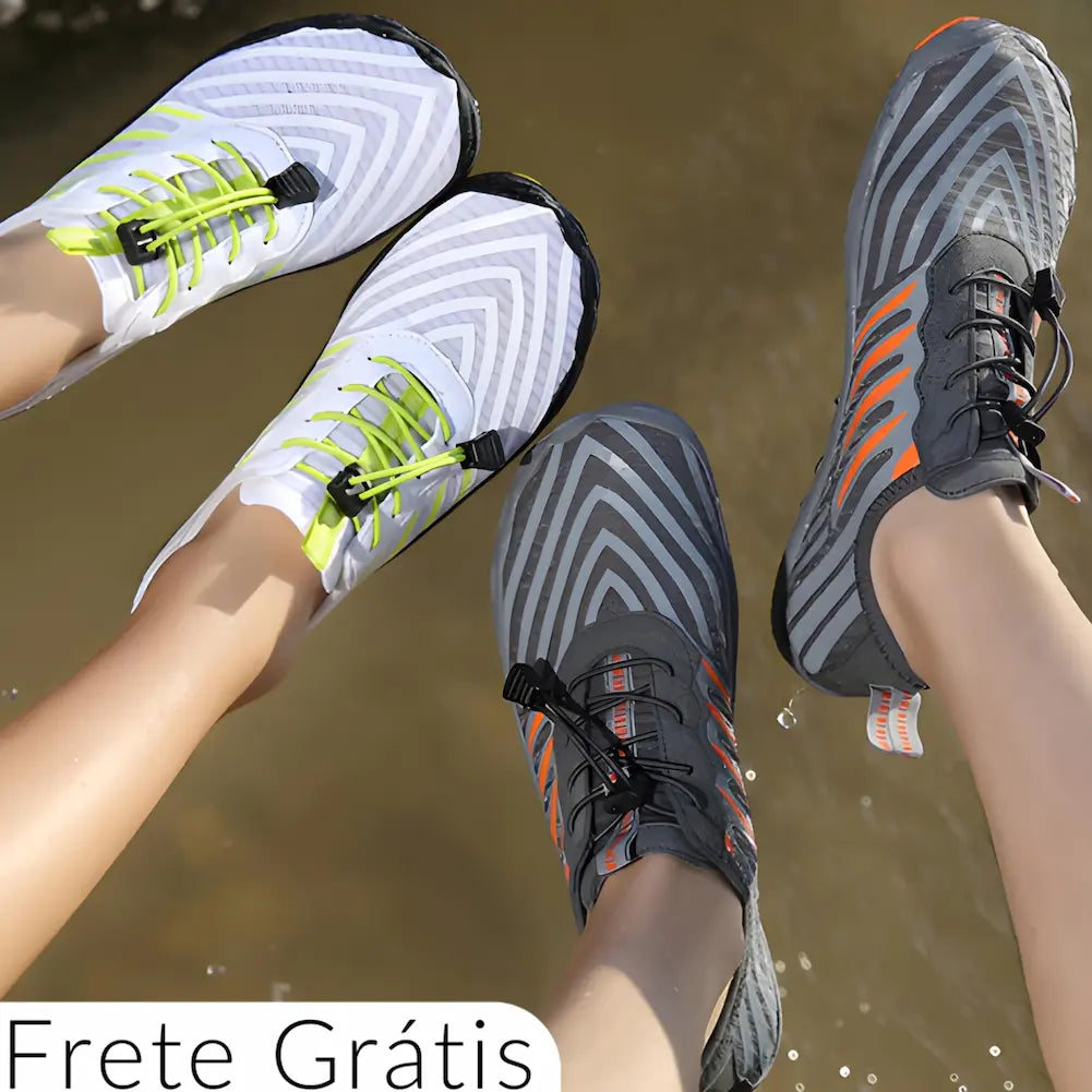 A imagem mostra dois pares de tênis esportivos, um branco com detalhes verdes e outro cinza com detalhes laranja, ambos com um design moderno e padrões em ziguezague. Eles estão em um ambiente ao ar livre, com água ao fundo, sugerindo que são adequados para atividades aquáticas ou trilhas. Os tênis possuem cordões elásticos e uma etiqueta indicando "Frete Grátis" na parte inferior esquerda. A presença de água sugere que os tênis têm boa secagem rápida, ideais para ambientes úmidos.