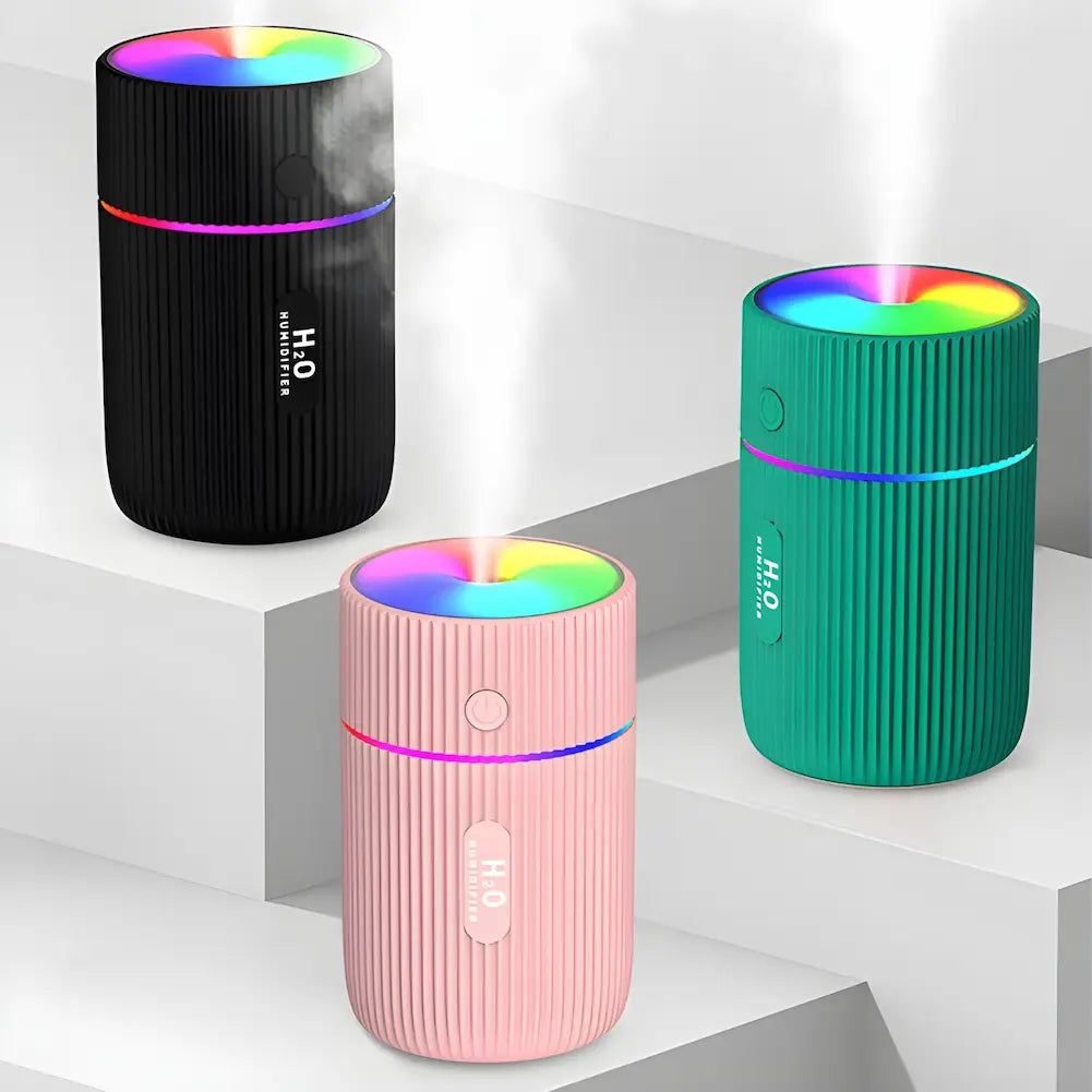 Imagem mostrando três umidificadores de ar portáteis, com design cilíndrico e textura ondulada, posicionados em diferentes níveis de uma estrutura em degraus. O umidificador preto está no topo, emitindo luzes coloridas e vapor. O umidificador verde está à direita, também emitindo luzes coloridas e vapor. O umidificador rosa está à frente, mais abaixo, emitindo luzes coloridas e vapor. Todos os umidificadores têm um anel luminoso ao redor do meio do aparelho.